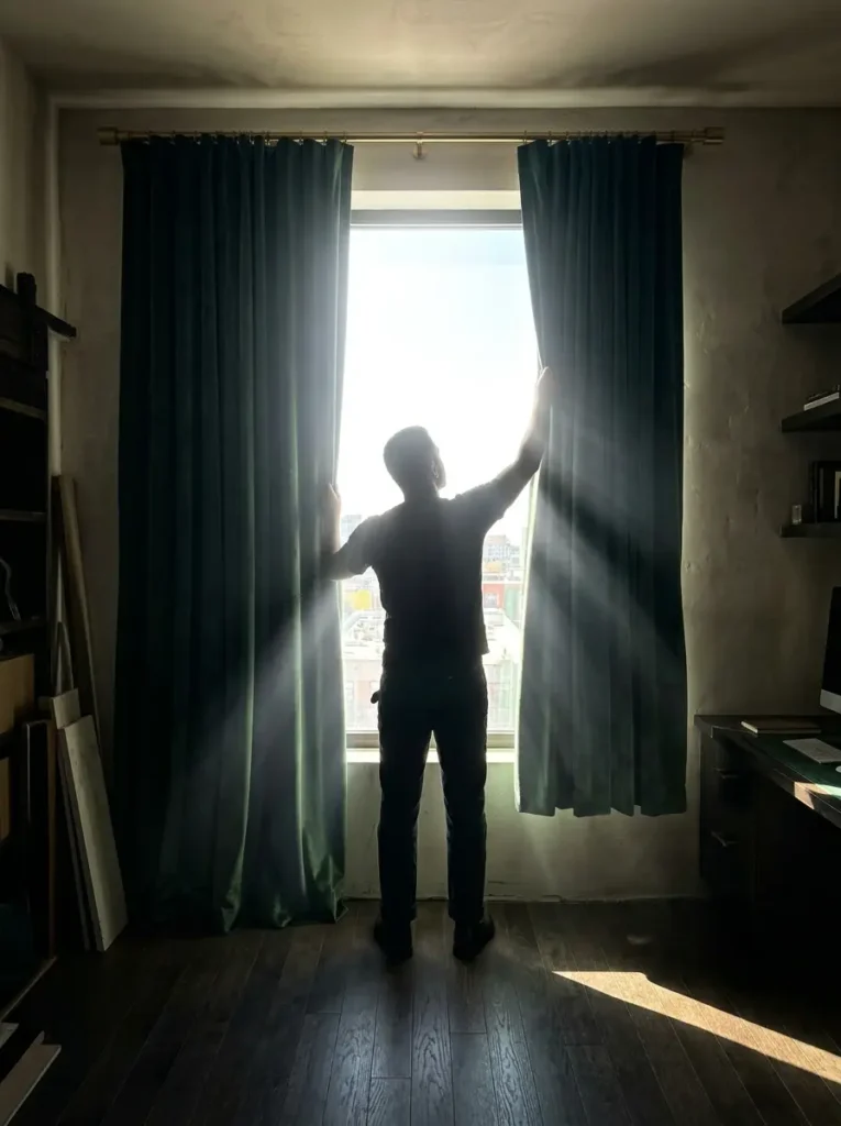 A person opens thick green velvet curtains revealing bright morning sunlight.