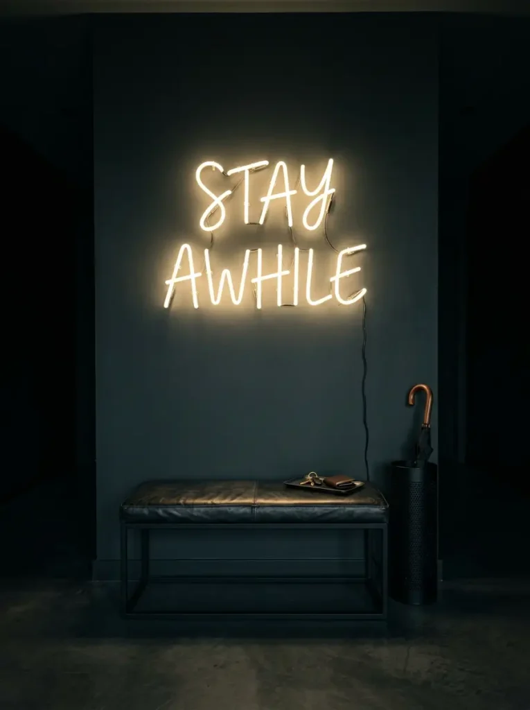 Glowing yellow neon sign reading Stay Awhile hangs above a black leather bench in a dark corridor.