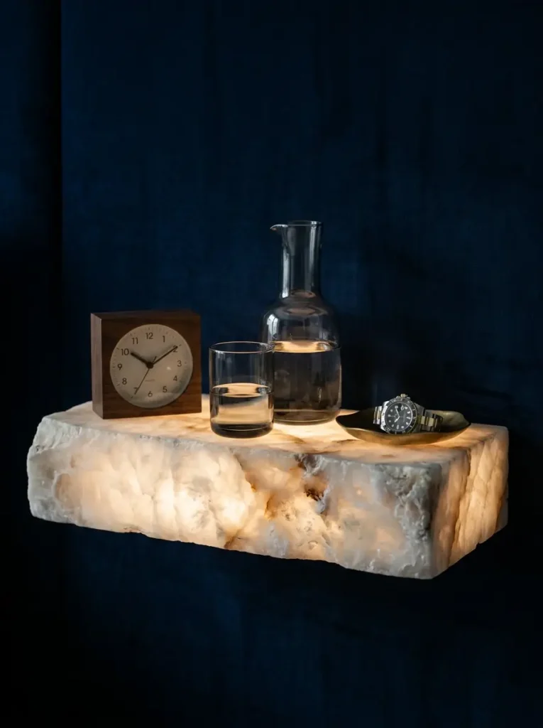 Glowing translucent stone floating shelf holding a clock and water pitcher next to a bed.