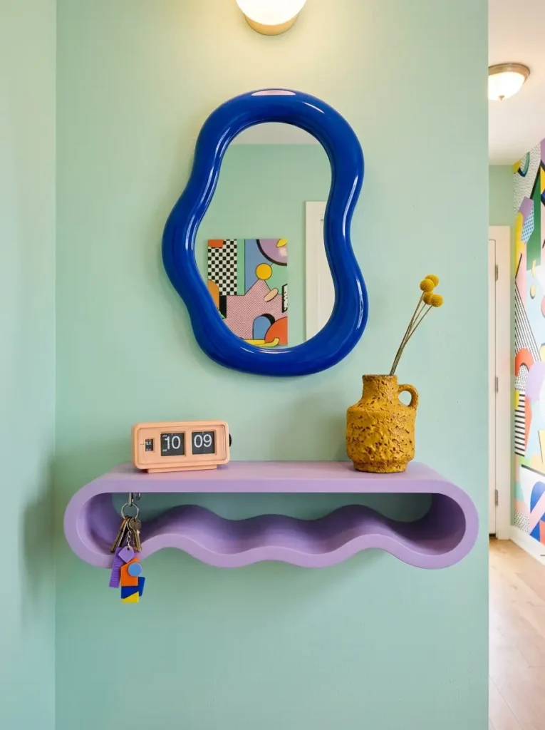 Wavy solid purple floating shelf holding keys and a yellow vase below an organic blue mirror.