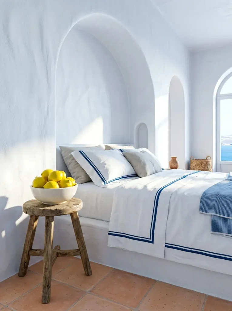 White stucco bedroom with deep arched walls crisp white sheets with navy blue borders and a wooden stool holding bright yellow lemons