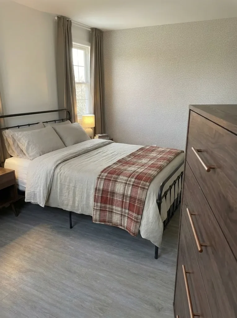 Elevated side view of a brick-red plaid quilt folded over flax bedding beside patterned walls and greige curtains.