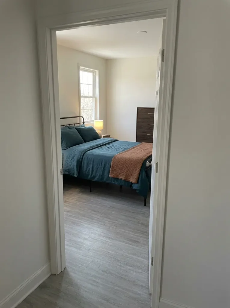 Deep-teal bedding and a copper-colored throw framed by an open doorway from an oyster-colored hallway.