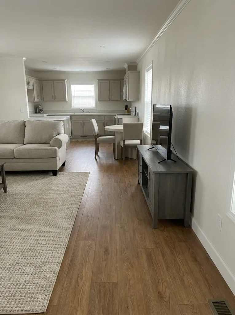 Open mobile home living room and kitchen where warm wood flooring provides the dominant hard-surface pattern and surrounding finishes stay quieter.
