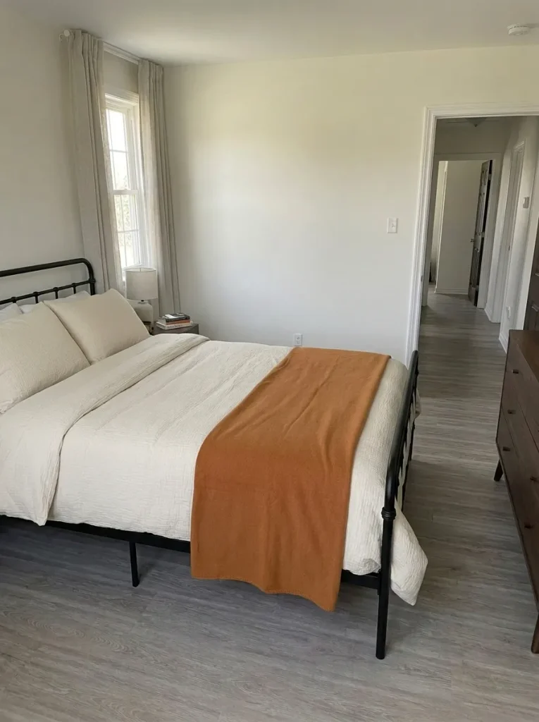 Low frontal view of a black metal bed frame with cream bedding and a pumpkin-colored throw.