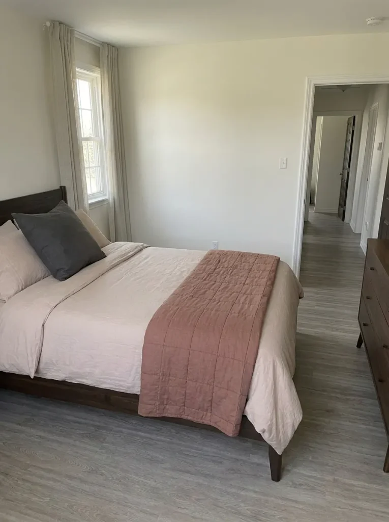 Putty bedding and a dusty-clay throw beside gray flooring extending through an open hallway doorway.
