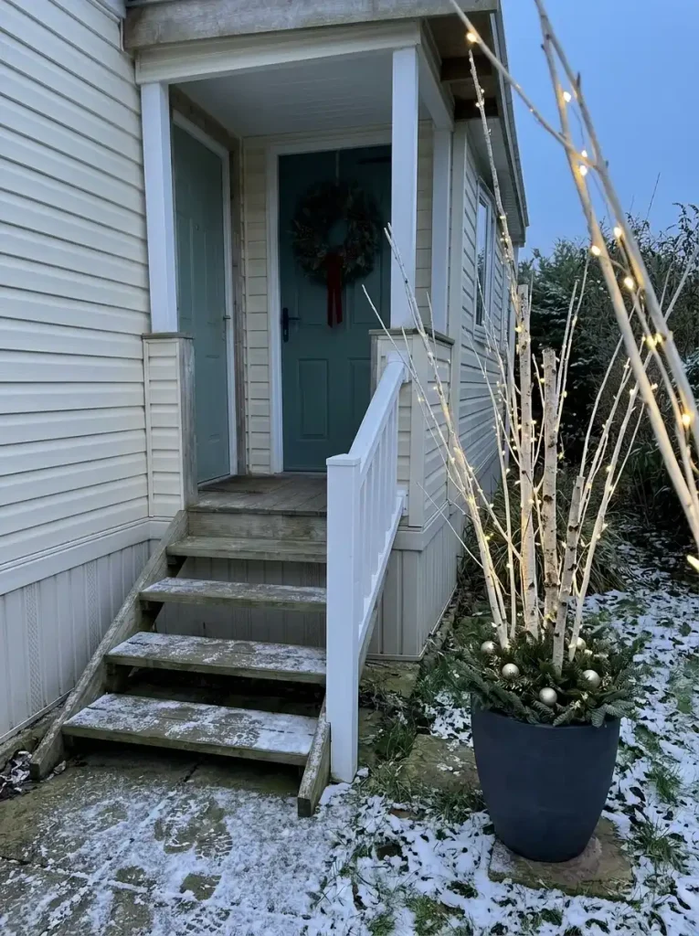 Illuminated birch branches in a gray planter beside a small mobile-home porch at dusk.