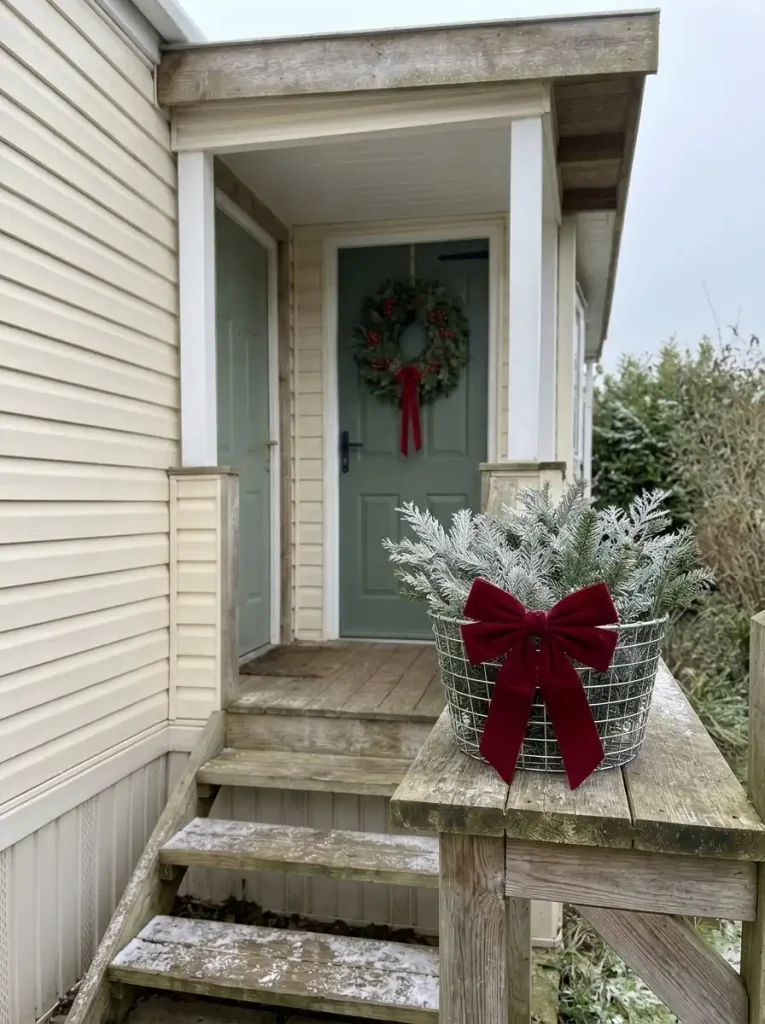 Galvanized wire basket filled with evergreen branches and a red velvet bow beside a mobile-home porch entrance.