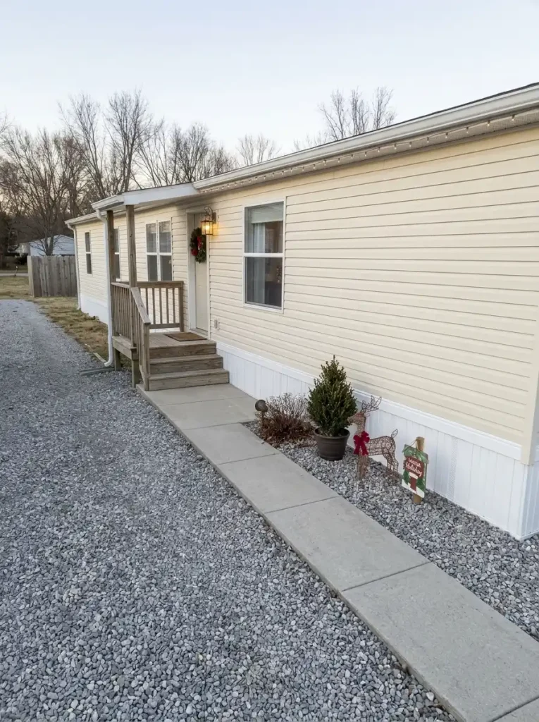 Small manufactured home with a gravel front yard and a compact Christmas display in an unused corner beside an open concrete walkway.