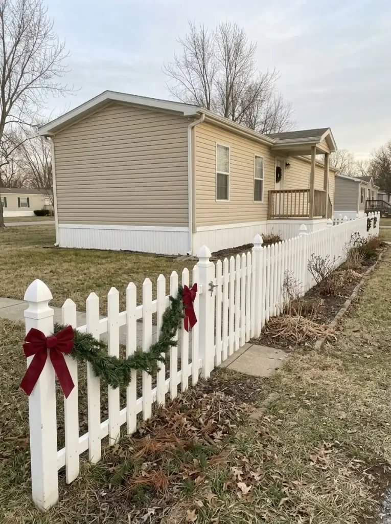Evergreen garland and red bows on one short white front-yard fence section beside a manufactured home, with its gate left clear.