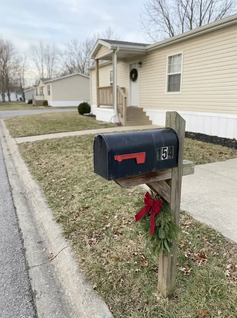 Curbside mailbox decorated with a small evergreen swag and red bow, with its flag and door visible and a modest mobile home behind it.