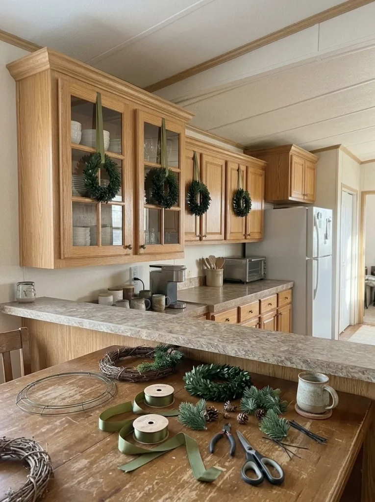 DIY mini Christmas wreath supplies on a dining table with finished wreaths hanging on mobile home kitchen cabinets.