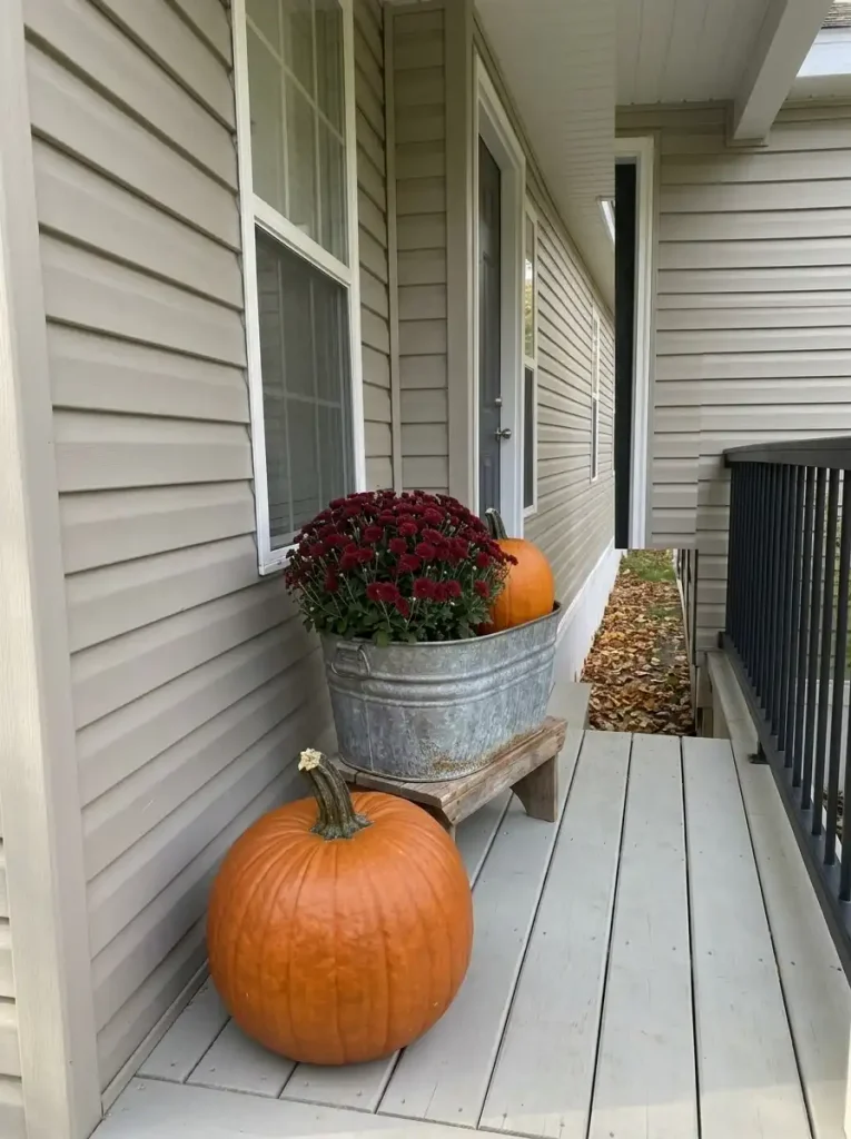 Orange pumpkin and burgundy mum raised on a galvanized pedestal beside a grounded pumpkin on a compact mobile-home porch.