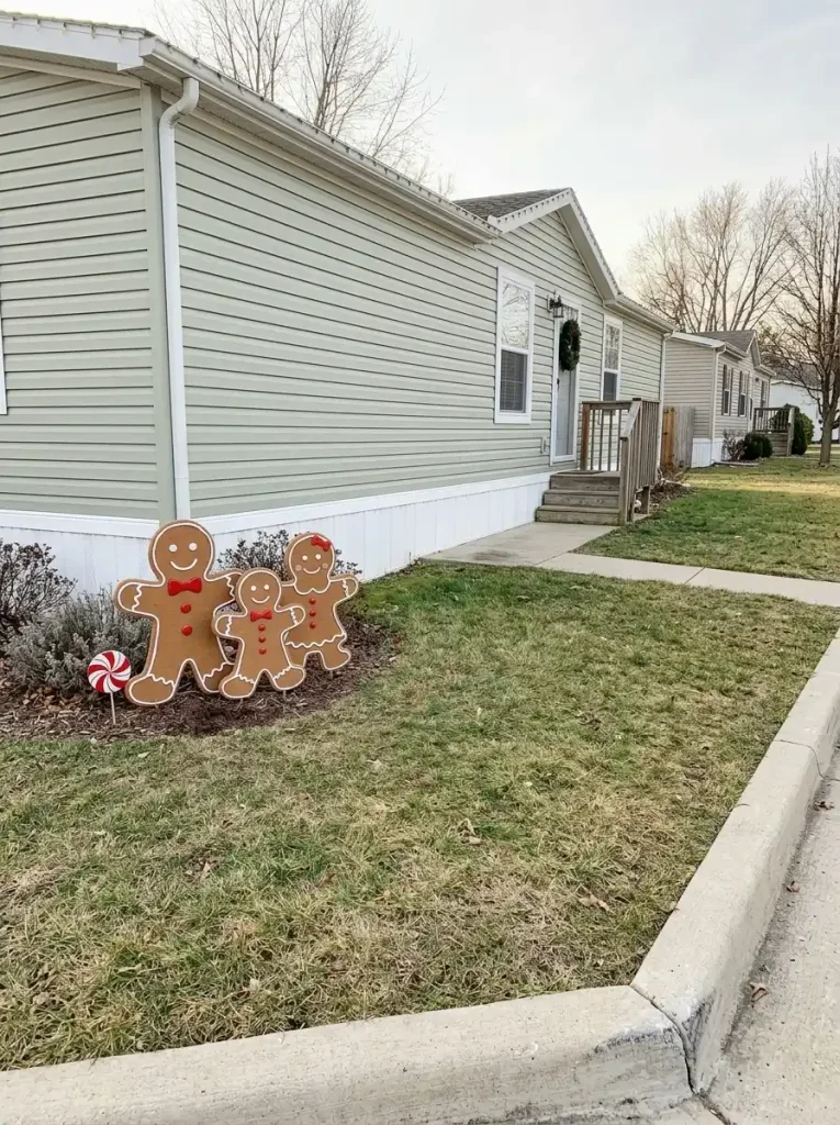 Three gingerbread character cutouts and a peppermint decoration grouped on one side of a small mobile-home lawn beside a clear entrance path.