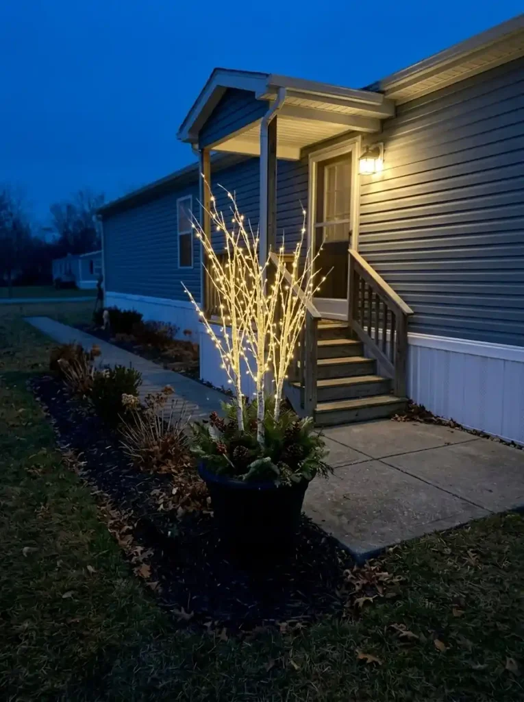 Warm-white illuminated birch-style branches in a planter on the edge of a small manufactured-home yard, with separate porch lighting and a clear path.