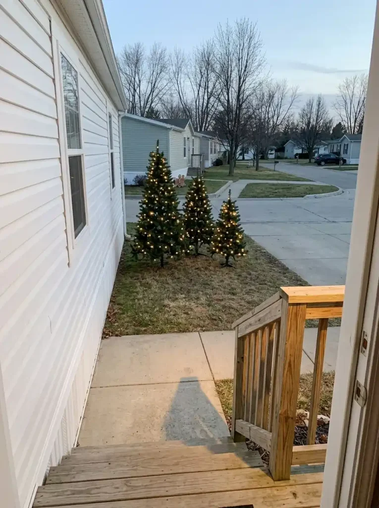 Three small Christmas trees arranged at different heights in an unused corner of a mobile-home yard, leaving the front path clear.