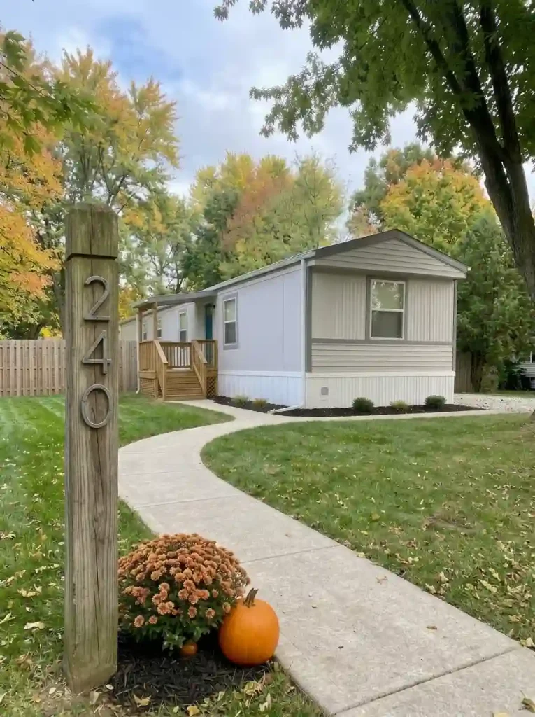 Low bronze mum and pumpkin below a readable address post, with the mobile home approach clear.