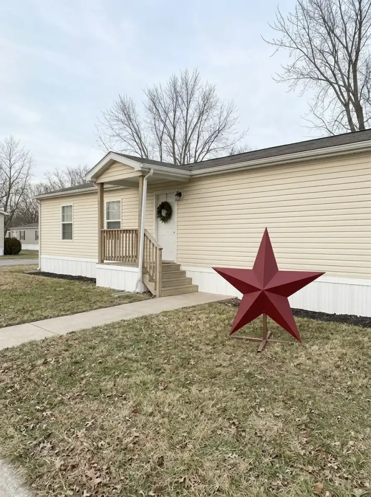Single oversized red Christmas star displayed on the lawn against plain siding, away from the windows and doorway of a small manufactured home.