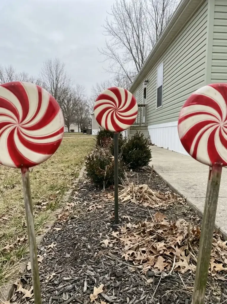 Three red-and-white peppermint disc decorations grouped in a narrow garden bed outside a pale-green single-wide mobile home.
