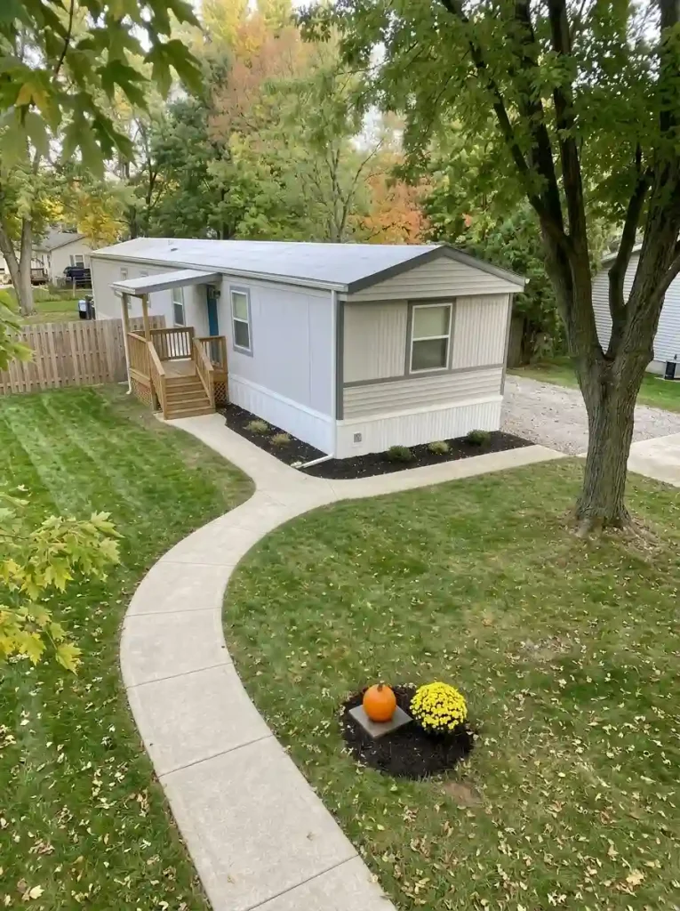 Orange pumpkin on a removable gray paver above damp mulch, beside a yellow mum in a mobile home front bed.
