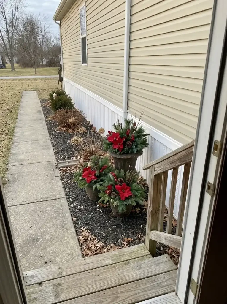 Three outdoor planters with red poinsettia-style foliage grouped beside a mobile-home entrance, outside the clear walking route.