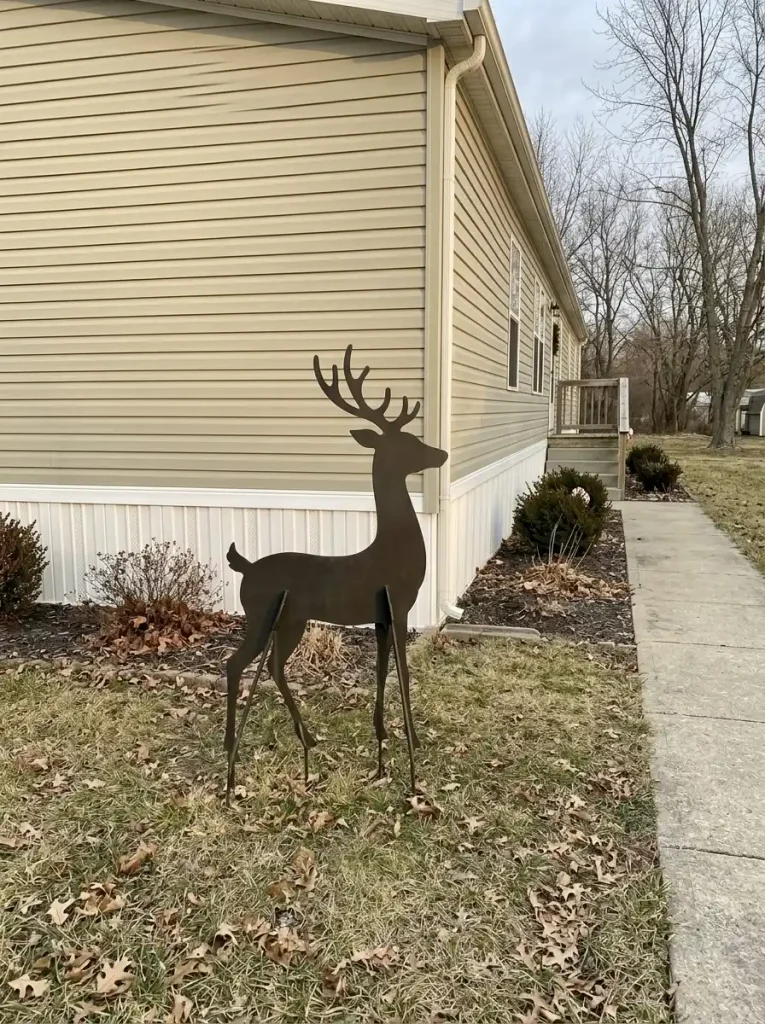 Dark reindeer silhouette standing in an open lawn pocket beside the light siding of a manufactured home, with an unobstructed walkway.