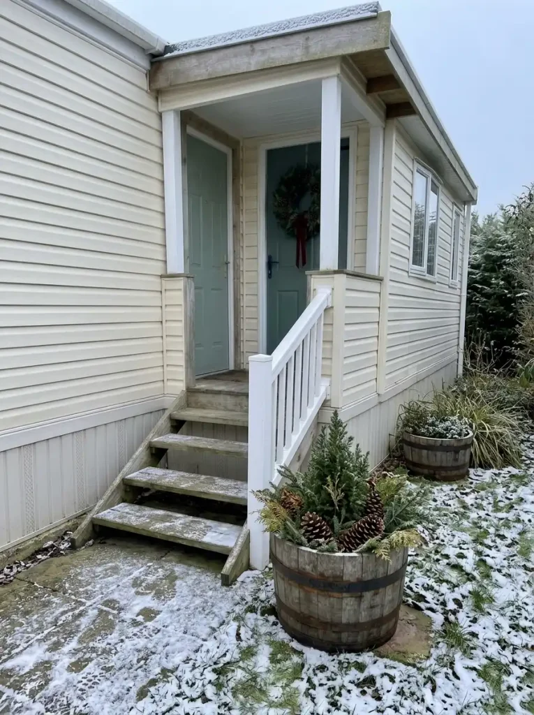 Two rustic wooden barrel Christmas planters with miniature evergreens and pinecones beside a manufactured-home entrance.
