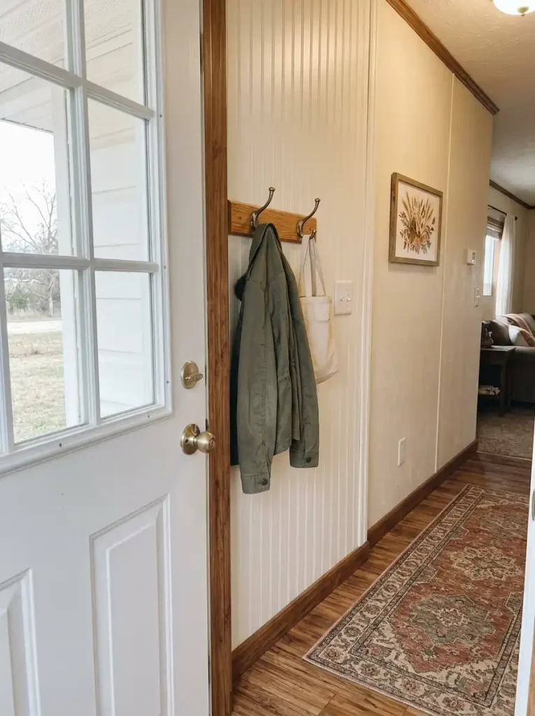 Short wooden peg rail beside an open mobile home door with a coat and tote hanging clear of the light switch.