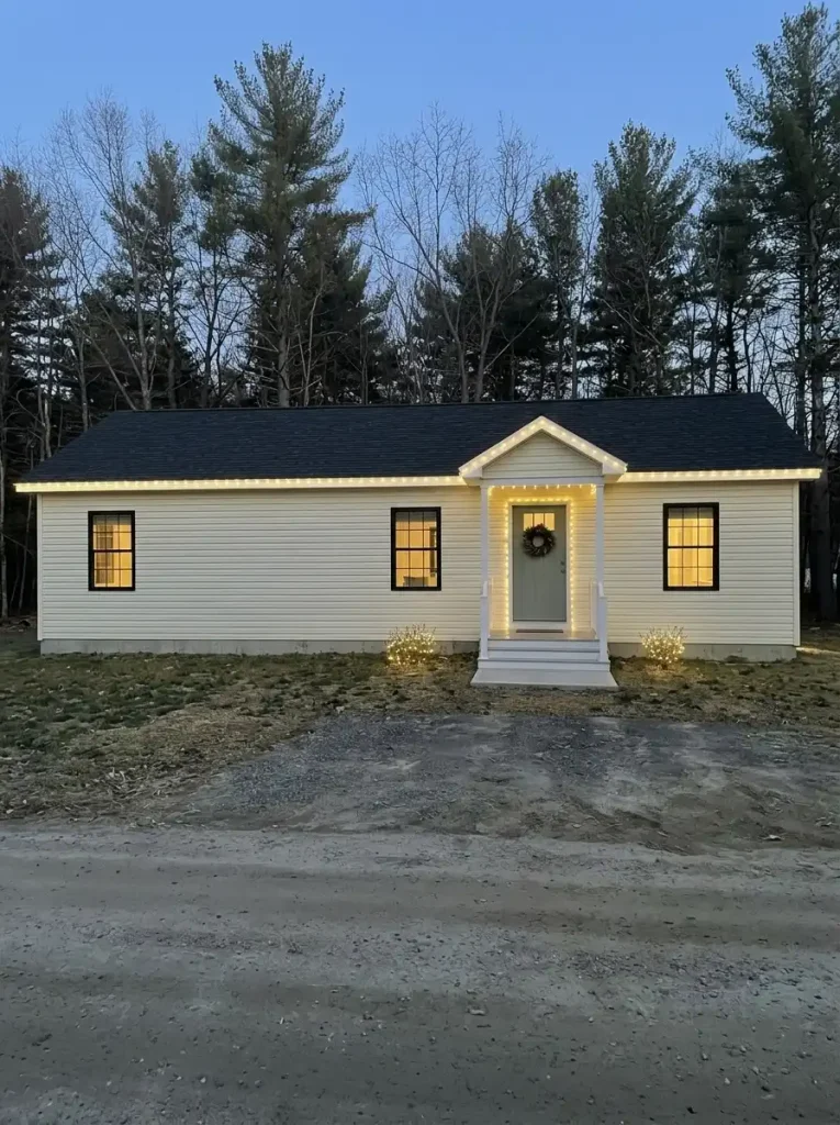 Finished outdoor Christmas lighting display on a cream single-wide manufactured home, photographed from the street at blue hour.