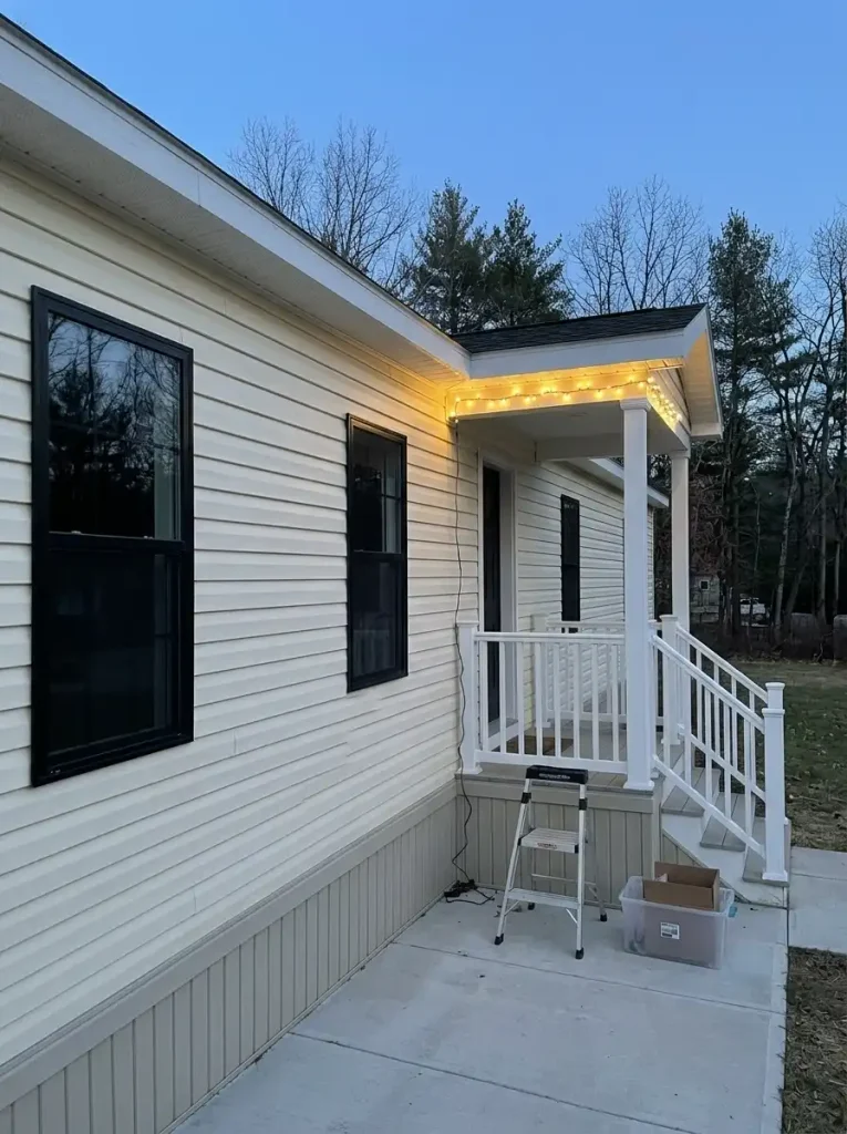 Warm-white outdoor LED Christmas lights tested along the covered porch of a cream single-wide mobile home at early dusk.