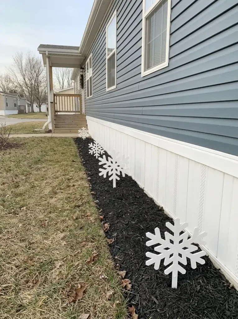 White snowflake decorations staggered in a narrow dark mulch strip along the blue-gray siding of a single-wide mobile home.
